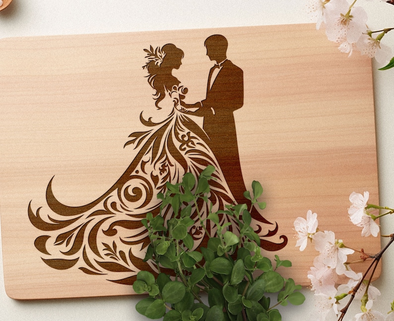 Pu&ograve; includere: Un tagliere in legno con un design a laser di una sposa e uno sposo. La sposa indossa un abito fluido con intricati motivi floreali e lo sposo &egrave; in piedi accanto a lei con il braccio intorno alla sua vita.