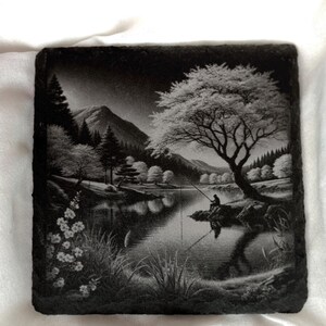 Puede incluir: Una baldosa de pizarra grabada en blanco y negro que representa una escena serena junto a un lago. Una figura solitaria se sienta en una roca junto a la orilla del agua, pescando con una ca&ntilde;a. Se pueden ver &aacute;rboles y una monta&ntilde;a en el fondo, creando una atm&oacute;sfera pac&iacute;fica y tranquila.