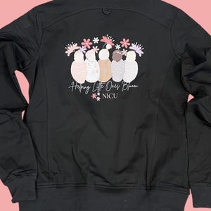 Puede incluir: Chaqueta negra con cremallera con un gráfico de cinco figuras estilizadas con flores en la cabeza. El texto "Helping Little Ones Bloom" y "**NICU" está impreso debajo de las figuras.