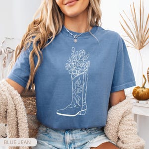 Puede incluir: Camiseta azul vaquero con un gr&aacute;fico blanco de una bota de vaquero llena de flores. La camiseta tiene mangas cortas y cuello redondo. El texto "BLUE JEAN" est&aacute; en la parte inferior de la imagen.
