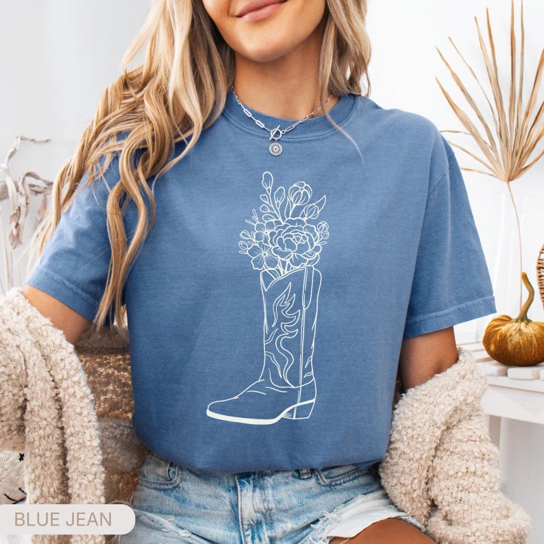 Puede incluir: Camiseta azul vaquero con un gr&aacute;fico blanco de una bota de vaquero llena de flores. La camiseta tiene mangas cortas y cuello redondo. El texto "BLUE JEAN" est&aacute; en la parte inferior de la imagen.
