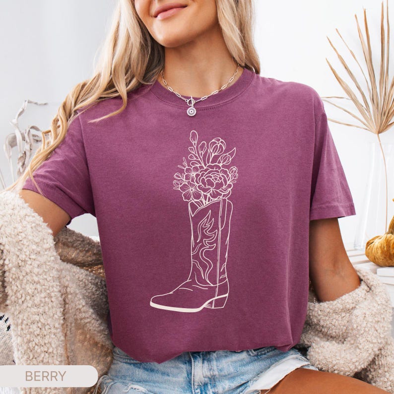 Puede incluir: Camiseta color bayas con un gr&aacute;fico blanco de una bota de vaquero llena de flores. La camiseta tiene mangas cortas y cuello redondo. La palabra "BERRY" est&aacute; impresa en la parte inferior de la imagen.