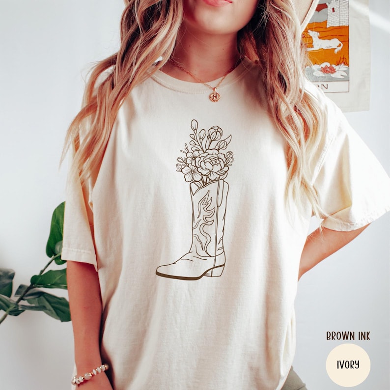 Puede incluir: Camiseta de color marfil con una ilustraci&oacute;n en tinta marr&oacute;n de una bota de vaquero llena de flores. La bota tiene costuras decorativas. La camiseta es de color neutro y tiene mangas cortas.