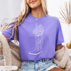 Puede incluir: Camiseta violeta con un gr&aacute;fico blanco de una bota de vaquero llena de flores. La palabra "VIOLET" est&aacute; impresa en blanco sobre un rect&aacute;ngulo blanco en la parte inferior de la imagen. La camiseta es de manga corta.