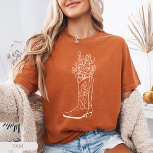Puede incluir: Camiseta de color naranja quemado con un gr&aacute;fico blanco de una bota de vaquero llena de flores. La bota tiene costuras decorativas. La camiseta es de manga corta y tiene un corte holgado. La palabra "YAM" es visible en la parte inferior izquierda.