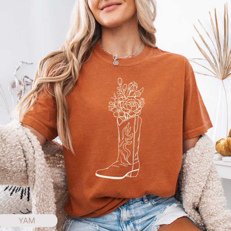Puede incluir: Camiseta de color naranja quemado con un gr&aacute;fico blanco de una bota de vaquero llena de flores. La bota tiene costuras decorativas. La camiseta es de manga corta y tiene un corte holgado. La palabra "YAM" es visible en la parte inferior izquierda.