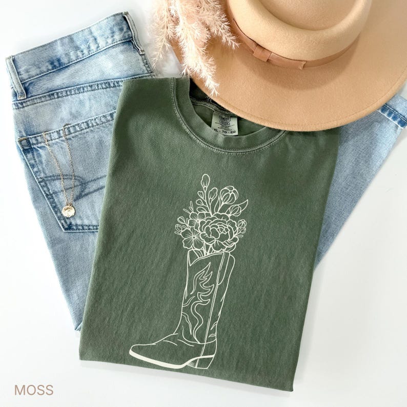 Puede incluir: Una camiseta verde musgo con un gr&aacute;fico blanco de una bota de vaquero llena de flores. La camiseta est&aacute; colocada sobre unos vaqueros azul claro, un sombrero beige y un collar dorado.