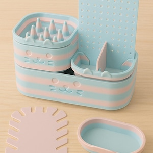 May include: A cat-themed grooming kit in pastel blue and pink. The kit includes a base with a cat face design, a grooming tool, a small tray, and a perforated panel. The set is designed for pet grooming.