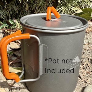 May include: A gray titanium camping pot with an orange handle and lid. The pot has a handle and a lid with an orange handle. The text "*Pot not Included" is printed on the side. The pot is designed for outdoor use.
