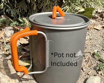 Può includere: Una pentola da campeggio in titanio grigio con manico e coperchio arancione. La pentola ha un manico e un coperchio con manico arancione. Il testo "*Pot not Included" è stampato sul lato. Progettata per uso esterno.