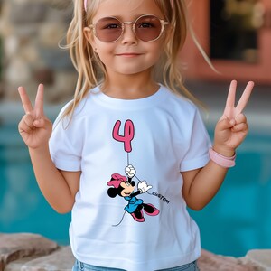 May include: White t-shirt featuring a pink number 4 balloon and a cartoon Minnie Mouse character in a blue dress, holding the balloon string. The word "custom" is printed on the shirt. The girl is wearing sunglasses and a pink headband.