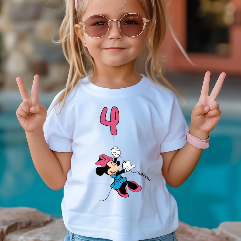 May include: White t-shirt featuring a pink number 4 balloon and a cartoon Minnie Mouse character in a blue dress, holding the balloon string. The word "custom" is printed on the shirt. The girl is wearing sunglasses and a pink headband.