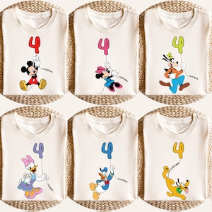 May include: Six cream-colored t-shirts featuring cartoon characters holding a number 4 balloon. Mickey Mouse, Minnie Mouse, Goofy, Daisy Duck, Donald Duck, and Pluto are depicted. Each shirt has the word "custom" printed on it.