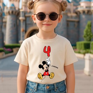 May include: A beige t-shirt featuring a Mickey Mouse graphic holding a red number 4 balloon. The word "custom" is printed next to Mickey. The shirt is worn by a young girl wearing sunglasses.