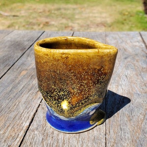 May include: A small, handmade ceramic cup with a speckled brown and yellow glaze. The cup has a blue base and a unique, asymmetrical shape.