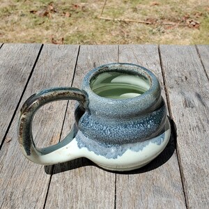 May include: A handmade ceramic mug with a blue and white glaze. The mug has a unique, wavy design and a wide, rounded handle.