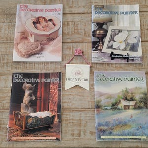 May include: Four issues of "The Decorative Painter" magazine are displayed on a wooden surface. The covers feature various decorative art subjects, including a heart-shaped box, a still life with a lamp, a cherub, and a landscape painting. A banner reading "Frozen in Time" hangs in the center.