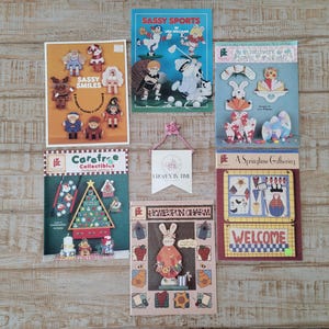 May include: A collection of craft pattern books and decorative items. The books feature various themes, including sports, holiday, and seasonal designs. The color palette includes blues, greens, reds, and yellows. The text "Sassy Smiles", "Sassy Sports", and "Welcome" are visible.