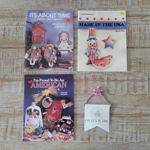 May include: Three craft pattern booklets titled It's About Time, Made in the USA, and I'm Proud To Be An American, alongside a small white pennant-shaped tag with a pink ribbon hanger and the text Frozen in Time Antiques printed in black.