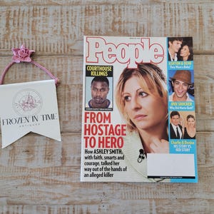 May include: A magazine cover of People magazine with the headline "From Hostage to Hero" next to a small banner with the text "Frozen in Time Antiques". The banner is white with a pink flower and pink rope.