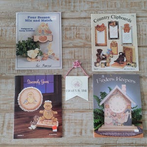 May include: An assortment of craft books and a decorative banner are displayed on a wooden surface. The books feature titles like "Four Season Mix and Match" and "Finders Keepers." The banner reads "Frozen in Time."