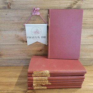May include: A stack of six vintage hardback books with maroon covers. The top book has the title "The Life and Letters of John Ruskin" printed on the spine. The books are sitting on a wooden surface.