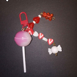 Op de afbeelding: Een sleutelhanger met een roze en witte lolly, rode hartkralen, aardbeienbedels en een witte snoepbedel. De sleutelhanger heeft een rode sluiting. Het nummer "1" staat in de linkerbovenhoek.