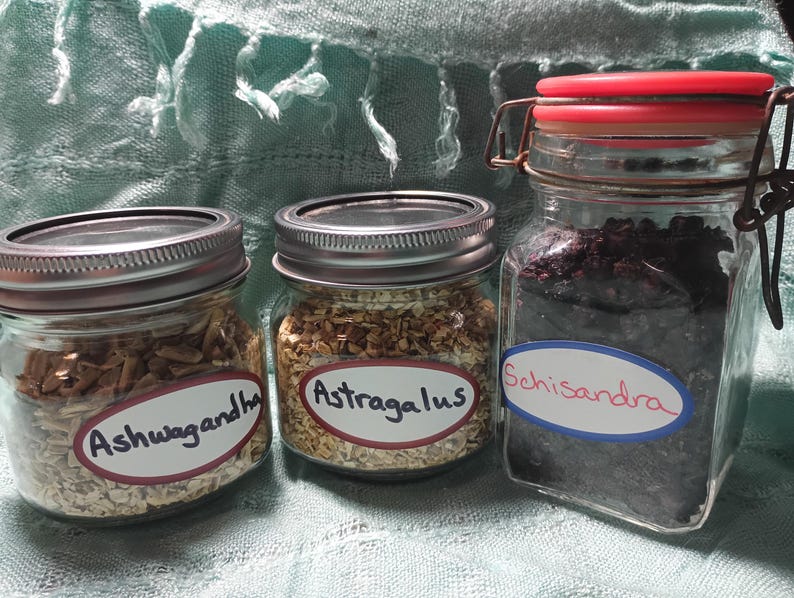 Puede incluir: Tres frascos de vidrio llenos de hierbas, etiquetados como "Ashwagandha", "Astragalus" y "Schisandra". Los frascos tienen tapas de metal y est&aacute;n sobre un fondo azul claro. El frasco de Schisandra tiene una tapa roja.