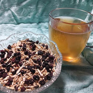 Puede incluir: Una taza de vidrio transparente llena de l&iacute;quido &aacute;mbar se encuentra junto a un cuenco de vidrio decorativo. El cuenco contiene una mezcla de bayas secas, palitos de color claro y otros ingredientes secos. El fondo es una tela azul claro texturizada.