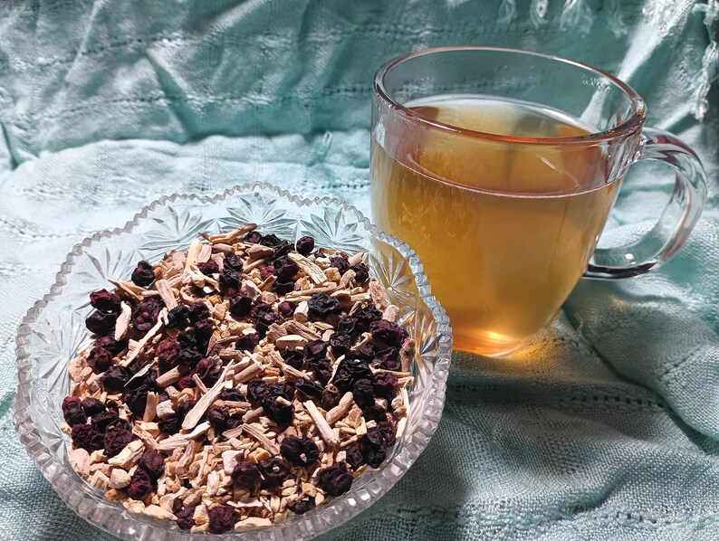 Puede incluir: Una taza de vidrio transparente llena de l&iacute;quido &aacute;mbar se encuentra junto a un cuenco de vidrio decorativo. El cuenco contiene una mezcla de bayas secas, palitos de color claro y otros ingredientes secos. El fondo es una tela azul claro texturizada.