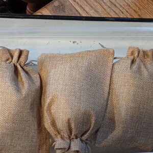 Puede incluir: Tres bolsas beige de yute con cord&oacute;n se colocan en fila. Las bolsas est&aacute;n hechas de una tela gruesa y tejida y tienen una parte superior fruncida. Las bolsas est&aacute;n dentro de un recipiente de pl&aacute;stico transparente.