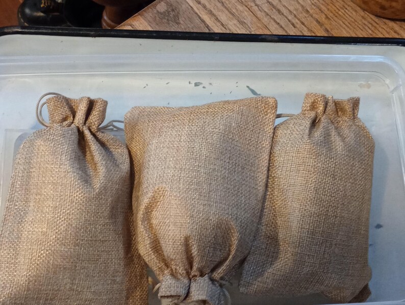 Puede incluir: Tres bolsas beige de yute con cord&oacute;n se colocan en fila. Las bolsas est&aacute;n hechas de una tela gruesa y tejida y tienen una parte superior fruncida. Las bolsas est&aacute;n dentro de un recipiente de pl&aacute;stico transparente.