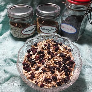 Puede incluir: Un cuenco de vidrio transparente lleno de una mezcla de hierbas y bayas secas. Tres frascos de vidrio con tapas de metal en el fondo, etiquetados como "Ashwagandha", "Astragalus" y "Schisandra". Los frascos contienen hierbas secas.