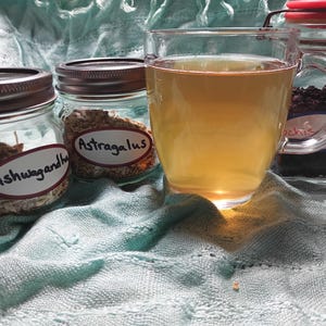 Puede incluir: Una taza de vidrio transparente llena de un l&iacute;quido &aacute;mbar claro se encuentra junto a tres frascos de vidrio. Dos frascos contienen hierbas etiquetadas como "Ashwagandha" y "Astragalus". El tercer frasco, con tapa roja, contiene hierbas oscuras. Sobre tela azul claro.
