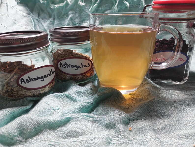 Puede incluir: Una taza de vidrio transparente llena de un l&iacute;quido &aacute;mbar claro se encuentra junto a tres frascos de vidrio. Dos frascos contienen hierbas etiquetadas como "Ashwagandha" y "Astragalus". El tercer frasco, con tapa roja, contiene hierbas oscuras. Sobre tela azul claro.