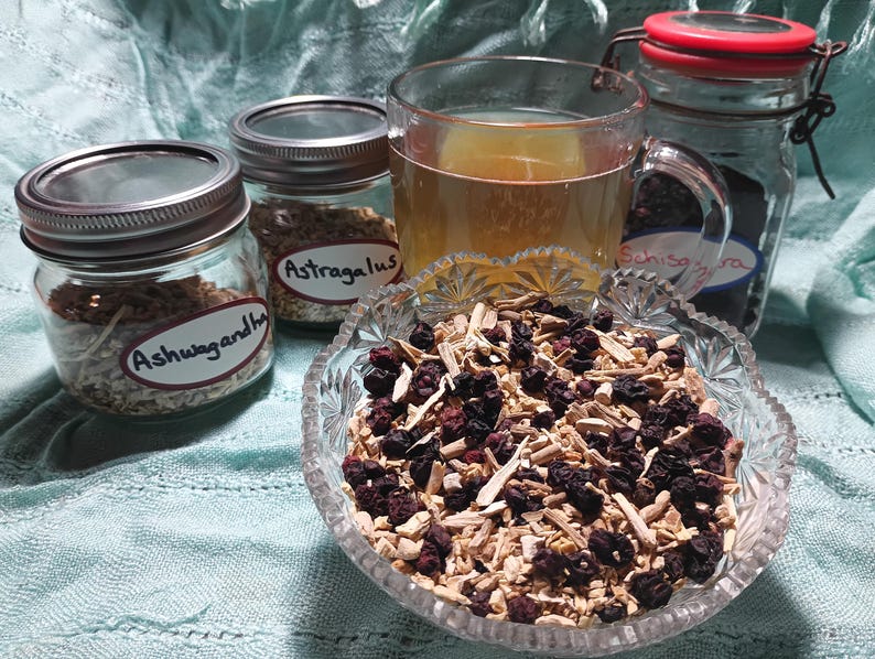 Puede incluir: Naturaleza muerta con ingredientes herbales para t&eacute;. Un cuenco de vidrio contiene una mezcla de bayas y ra&iacute;ces secas. Se ven tres frascos de vidrio etiquetados "Ashwagandha", "Astragalus" y "Schisandra", junto con una taza de t&eacute;.