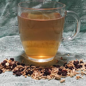 Puede incluir: Una taza de vidrio transparente llena de un l&iacute;quido marr&oacute;n claro, probablemente t&eacute; o una bebida similar. Un mont&oacute;n de hierbas y bayas secas se encuentra frente a la taza sobre una tela verde claro. La taza tiene un asa.