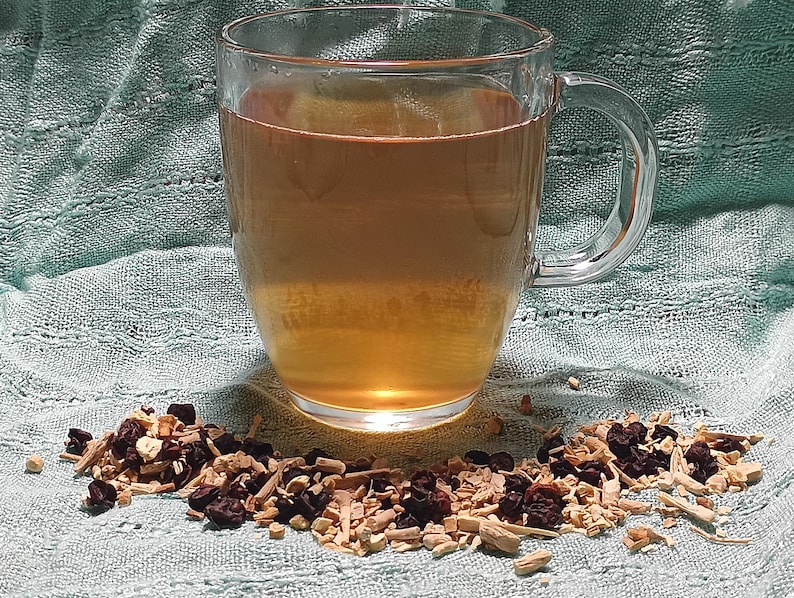 Puede incluir: Una taza de vidrio transparente llena de un l&iacute;quido marr&oacute;n claro, probablemente t&eacute; o una bebida similar. Un mont&oacute;n de hierbas y bayas secas se encuentra frente a la taza sobre una tela verde claro. La taza tiene un asa.