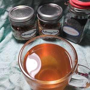 Puede incluir: Una taza de vidrio transparente llena de l&iacute;quido &aacute;mbar, probablemente t&eacute; o una infusi&oacute;n de hierbas, en primer plano. Detr&aacute;s, tres frascos de vidrio con tapas de metal, etiquetados "Ashwagandha", "Astragalus" y "Schisandra", sugiriendo remedios herbales.