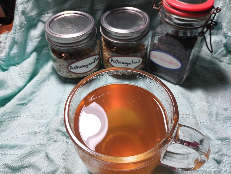 Puede incluir: Una taza de vidrio transparente llena de l&iacute;quido &aacute;mbar, probablemente t&eacute; o una infusi&oacute;n de hierbas, en primer plano. Detr&aacute;s, tres frascos de vidrio con tapas de metal, etiquetados "Ashwagandha", "Astragalus" y "Schisandra", sugiriendo remedios herbales.
