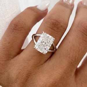 Op de afbeelding: Een verlovingsring met een radiant-geslepen diamant, voorzien van een grote, rechthoekige, gefacetteerde middensteen gezet in een gepolijste, zilverkleurige metalen band met een gespleten schacht die taps toeloopt naar de steen.