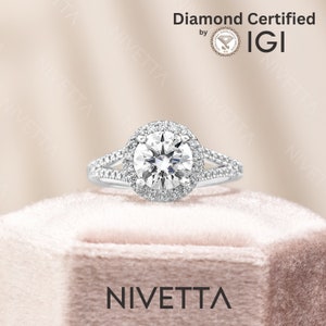 May include: A silver engagement ring with a round, clear diamond center stone surrounded by a halo of smaller diamonds. The ring has a split shank with two bands of smaller diamonds. The ring is in a white velvet box with the word "NIVETTA" printed on the front. The ring is certified by IGI.