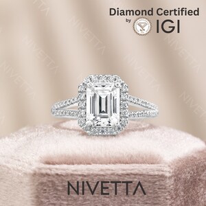 May include: A silver engagement ring with a large emerald cut diamond center stone surrounded by a halo of smaller diamonds. The ring has a split shank with pave diamonds. The ring is in a velvet box with the word "Nivetta" printed on the front. The ring is certified by IGI.
