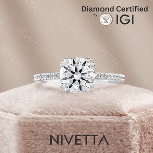 May include: A silver engagement ring with a round, clear diamond center stone and smaller diamonds set in the band. The ring is in a velvet box with the word "NIVETTA" printed on the front. The ring is certified by IGI.
