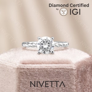 May include: A silver engagement ring with a round, clear diamond. The ring has a twisted band and is set in a box with the word "NIVETTA" printed on the front. The diamond is certified by IGI.