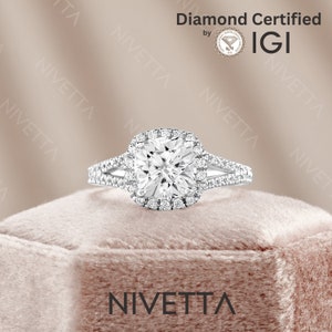 Op de afbeelding: Een zilveren ring met een vierkante diamant die in een halo-setting is gezet. De diamant is omgeven door kleinere diamanten. De ring heeft een gesplitste schacht met twee banden die in het midden samenkomen. De ring ligt op een roze fluwelen doos. De tekst "Diamond Certified by IGI" en "NIVETTA" is zichtbaar in de afbeelding.