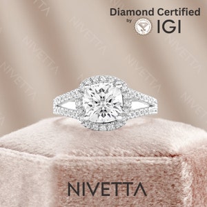 May include: A silver ring with a square-shaped diamond surrounded by a halo of smaller diamonds. The ring has a split shank with two bands. The ring is on a pink velvet box. The text "Diamond Certified by IGI" and "NIVETTA" is visible in the image.