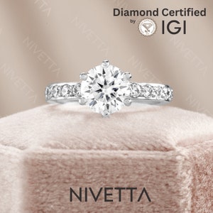 May include: A silver engagement ring with a round, clear diamond center stone and a band of smaller diamonds. The ring is in a velvet box with the word "Nivetta" printed on the front. The ring is certified by IGI.