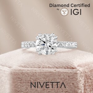May include: A diamond engagement ring with a round brilliant cut center stone and a pave diamond band. The ring is in a white gold setting and is displayed in a velvet box. The ring is certified by IGI.