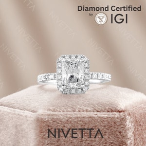 May include: A silver engagement ring with a large, rectangular, clear diamond center stone, surrounded by smaller diamonds. The ring band is also encrusted with small diamonds. The ring is set against a soft, pink background, with the word "NIVETTA" at the bottom.